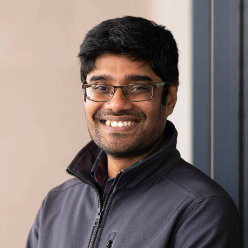 A man wearing glasses and a gray sweater smiles at a camera in front of a tan brown background.